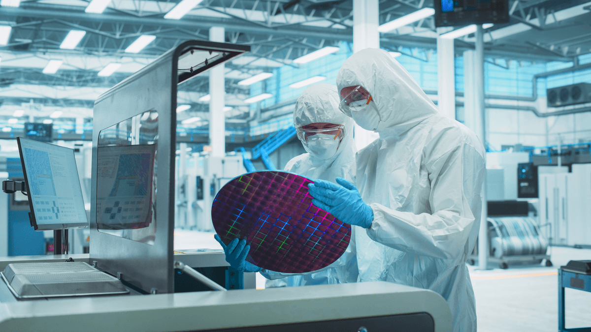Two semiconductor engineers in cleanroom suits examining a silicon wafer at a fabrication workstation